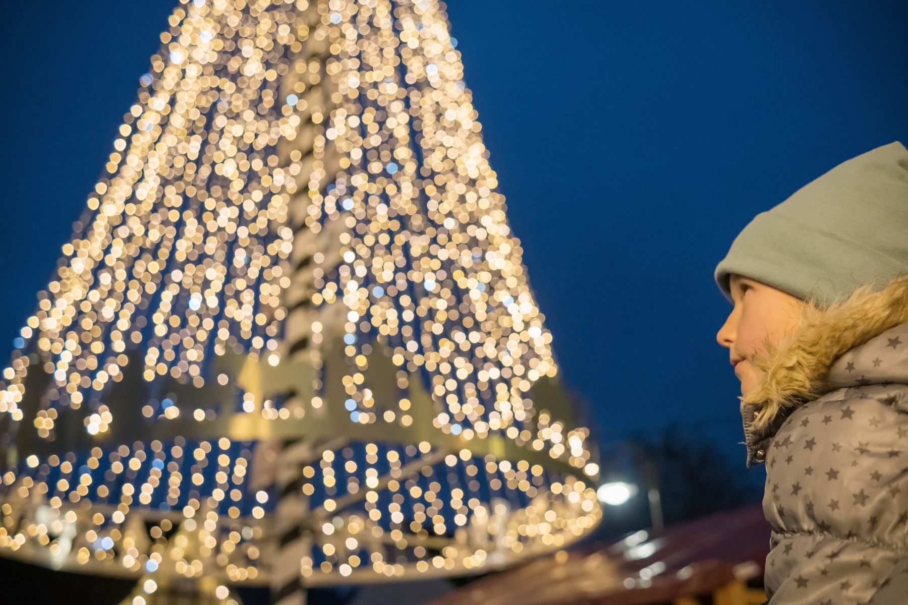Ein kleines Kind in grauem Wintermantel und Mütze blickt auf einen großen, hell erleuchteten und mit weißen Lichterketten geschmückten Weihnachtsbaum vor einem dunklen Abendhimmel.